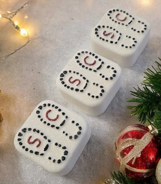 Square bath bombs with 'Santa's footprint' design, placed on a surface with Christmas decorations around, including lights and baubles