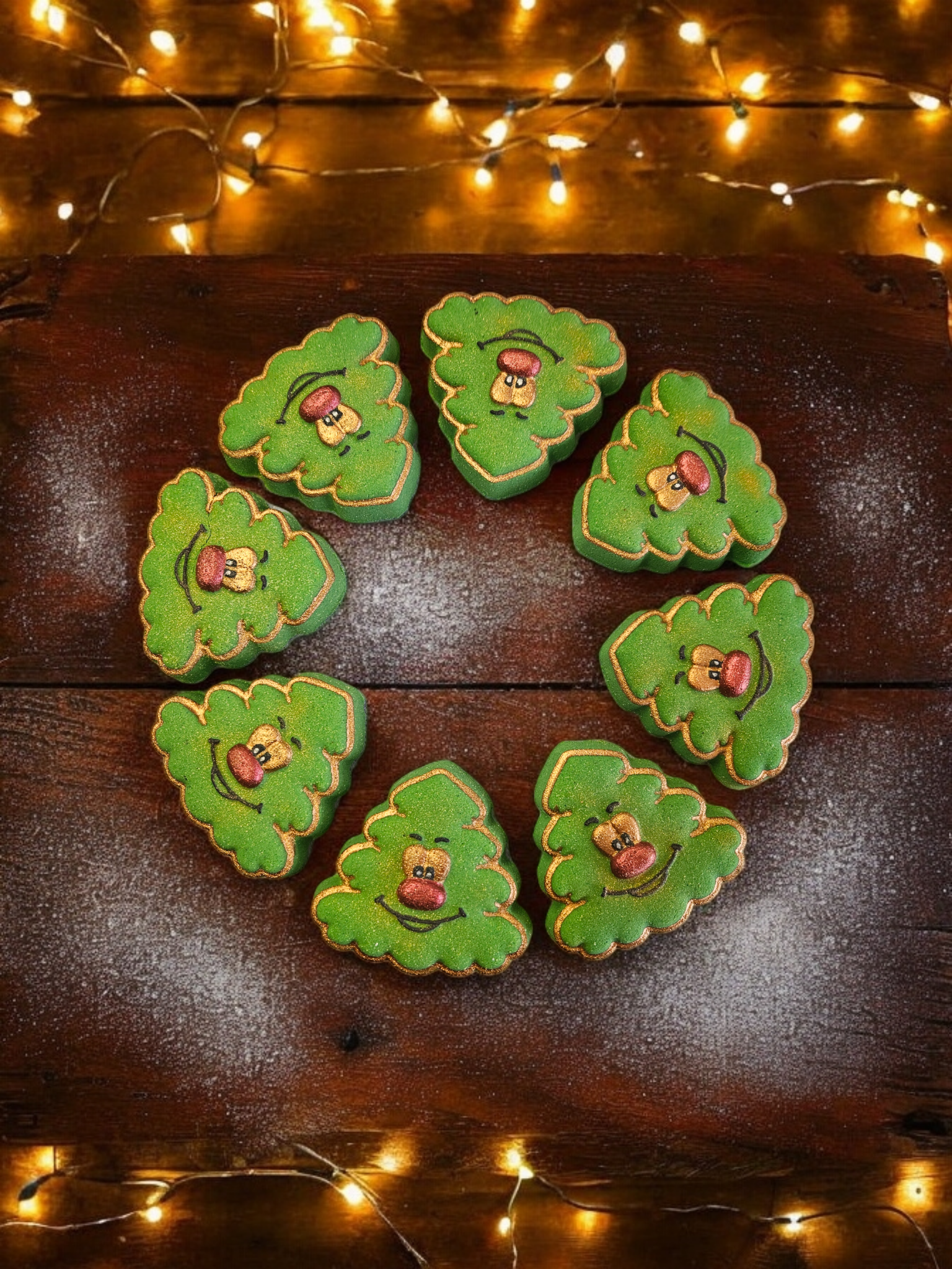 Green bath bomb Christmas tree shapes with a smiley face design, arranged in a circle presented on a dark surface with fairy lights in the background.